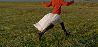 woman in red jumper and white long skirt and black boots running in a green landscape